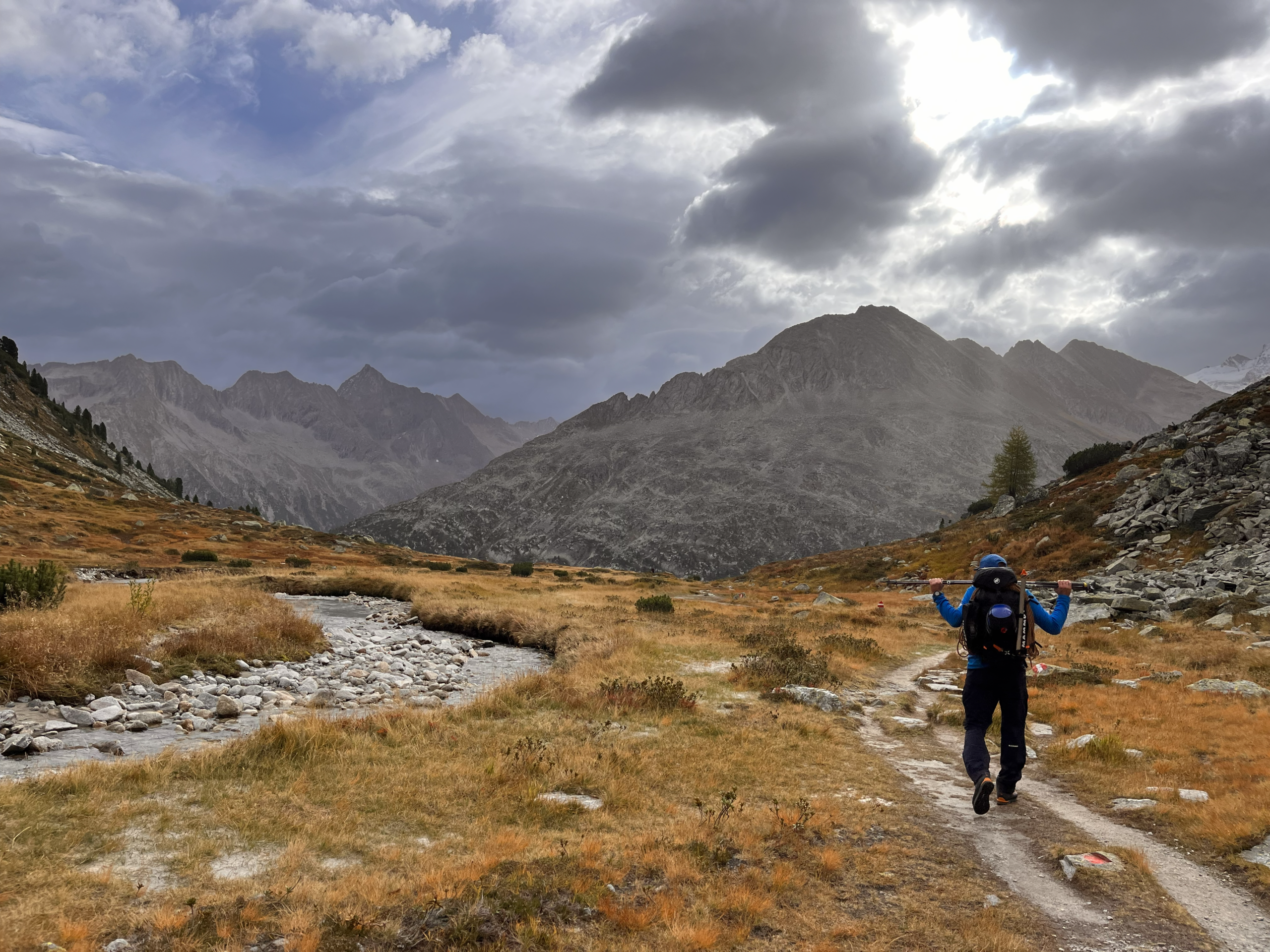 Bergkompass Bergwanderführer Pascal Schirmer als Bergsteiger mit großem Rucksack auf einem herbstlichen Bergpfad, umgeben von goldenen Gräsern und Felsen; im Hintergrund eine markante, felsige Bergkette unter dramatisch dunklen Wolken.