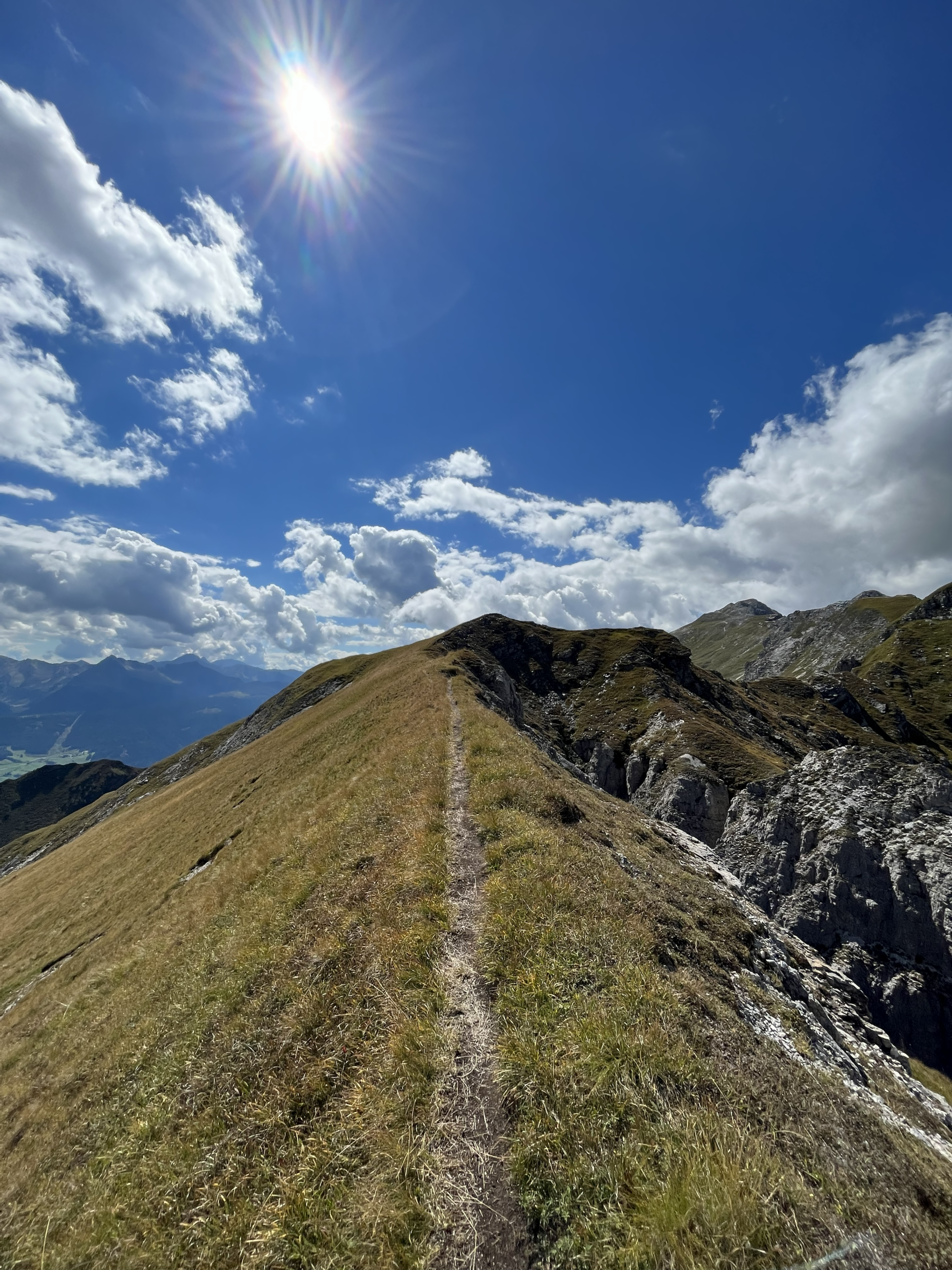 Schmaler Gratweg in Südtirol mit grasigen Rücken und steilen Felsflanken, der entlang des Bergkamms verläuft; im Hintergrund weite Bergketten und der Blick hinunter ins Tal - fotografiert von Bergkompass Bergwanderführer Pascal Schirmer