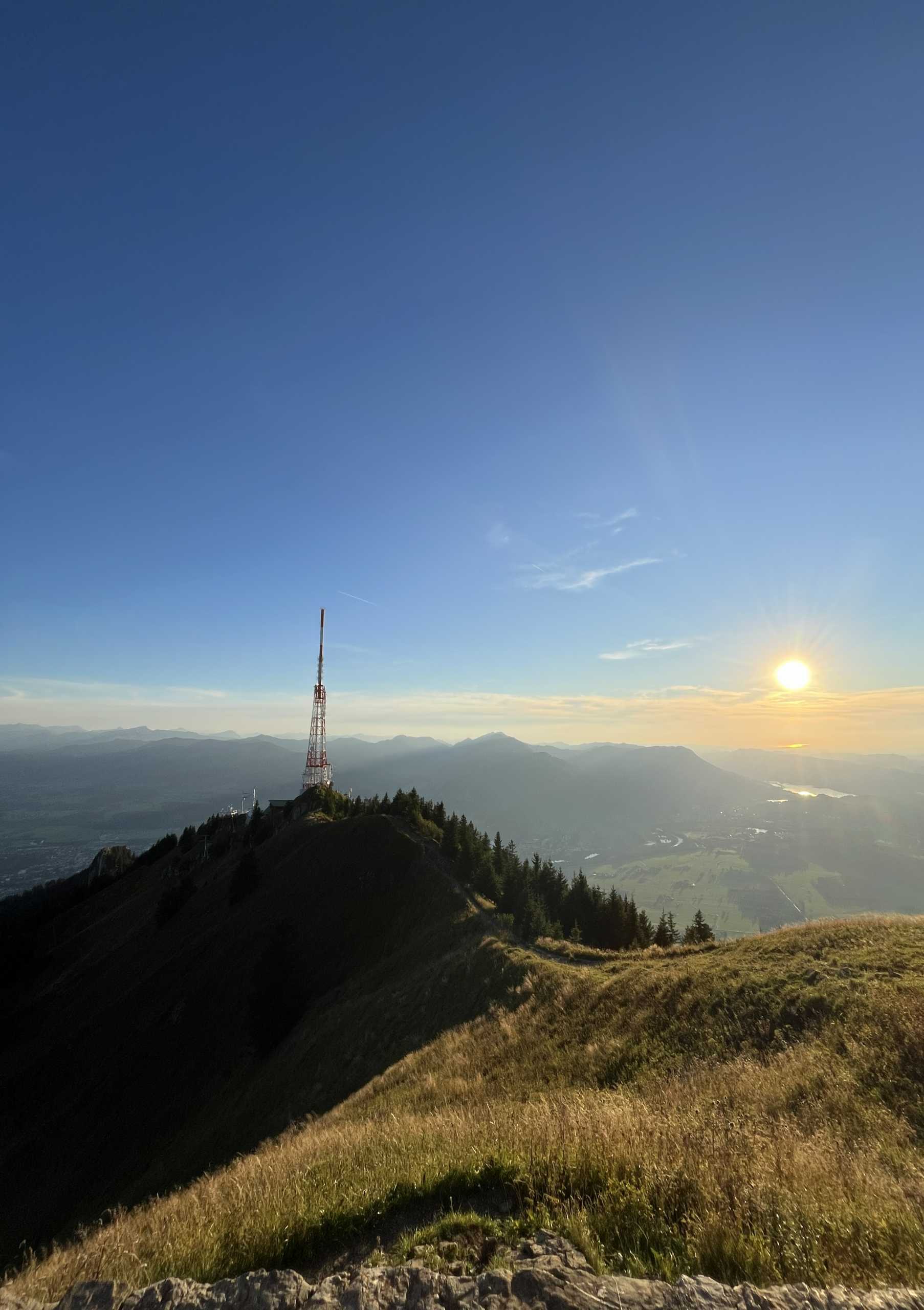 Blick vom Grünten auf den rot‑weißen Sendemast im Sonnenuntergang, mit Panorama über den Großen Alpsee, die Nagelfluhkette und in der Ferne den Bodensee – fotografiert von Bergkompass Bergwanderführer Pascal Schirmer