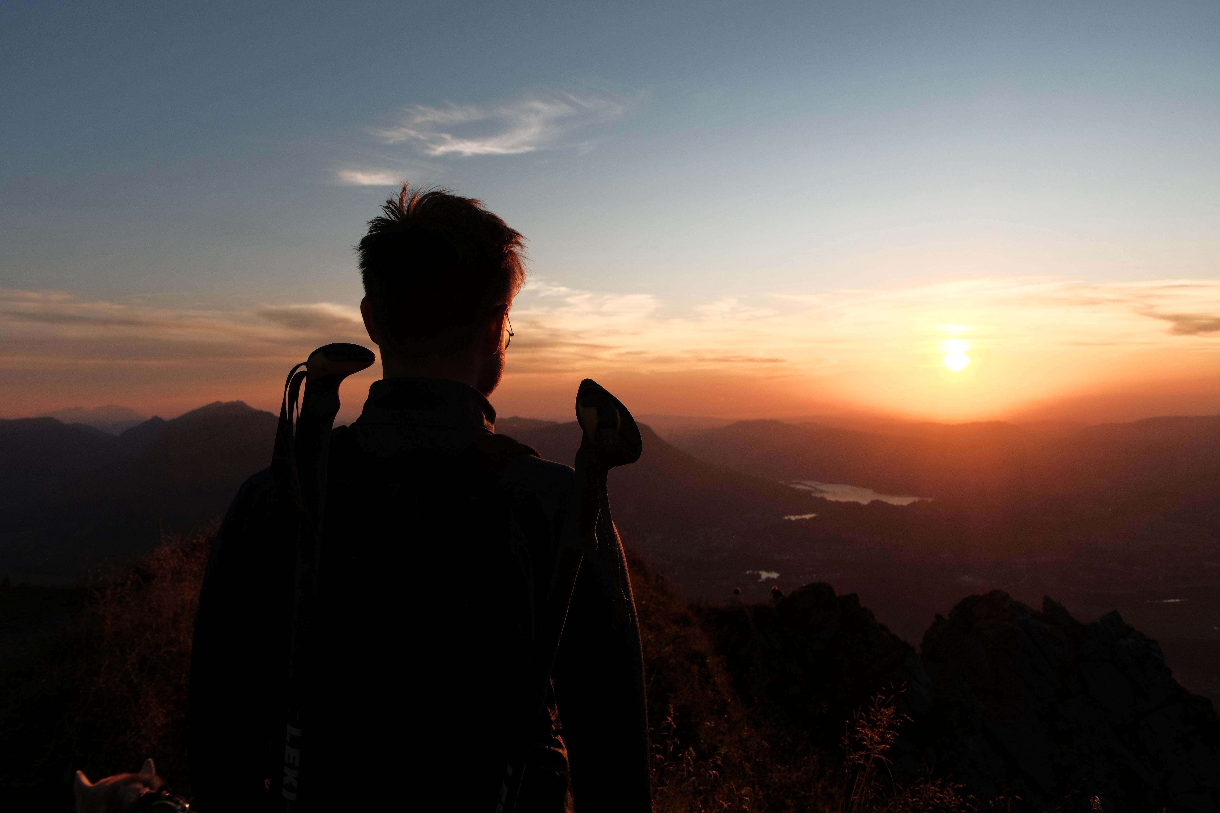 Wanderer am Grüntengipfel im Sonnenuntergang, Silhouette mit Stöcken vor orangefarbenem Himmel und Blick auf den Großen Alpsee im Tal – fotografiert von Bergkompass Bergwanderführer Pascal Schirmer