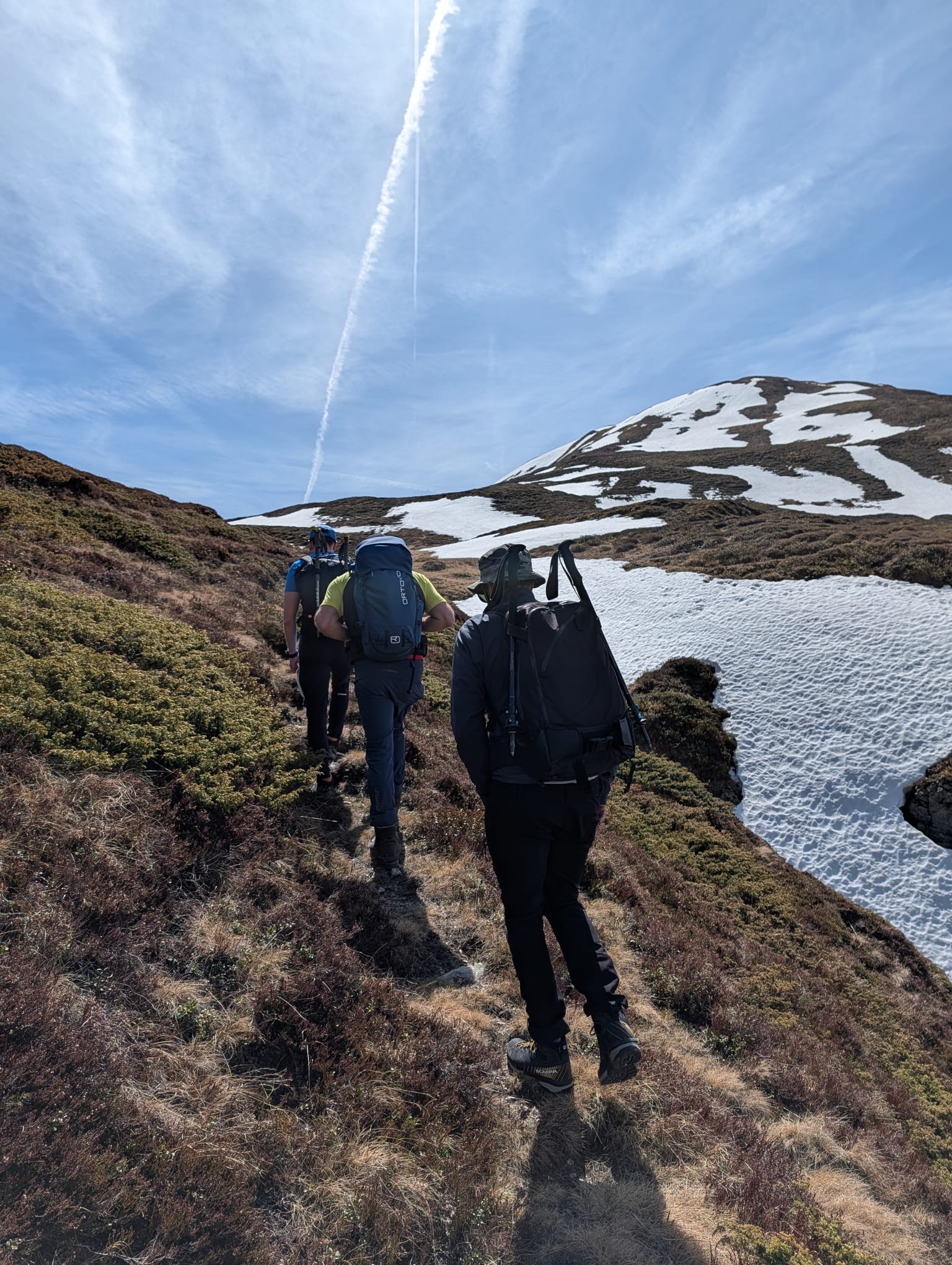Drei Wanderer mit Rucksäcken auf einem hochalpinen Pfad, Übergang von Schneefeldern zu trockenem Gras, weiter Blick zu schneebedeckten Gipfeln - Bergkompass Bergwanderführer Pascal Schirmer