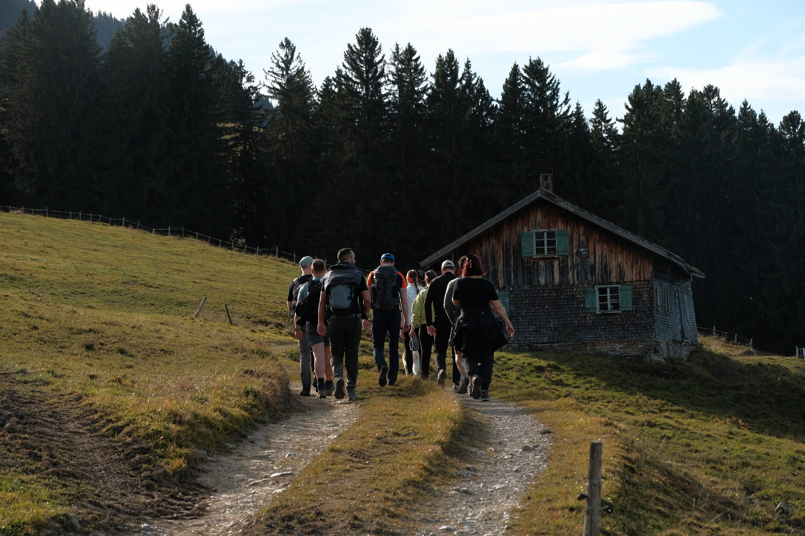 Wandernde Bergkopmass-Gruppe auf einem Almweg im Allgäu, der zu einer alten Holzhütte mit grünen Fensterläden führt; grasige Hanglage, dahinter dichter Bergwald und warmes Spätnachmittagslicht